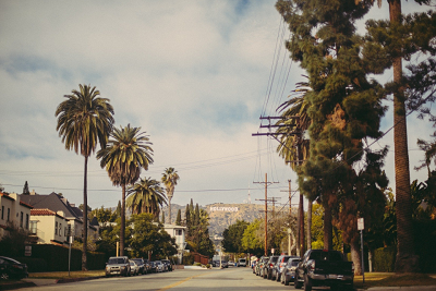 A view of a street lined with cars and palm trees, with the Hollywood sign on the hills in the background