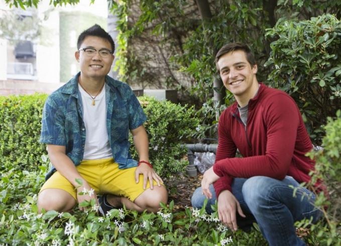 Two students posing for a photo in front of shrubs/trees.