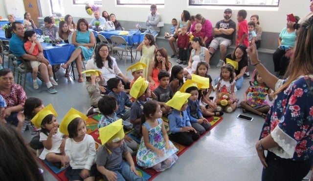A group of children sitting on the floor, while adults sit on tables and benches around the group.