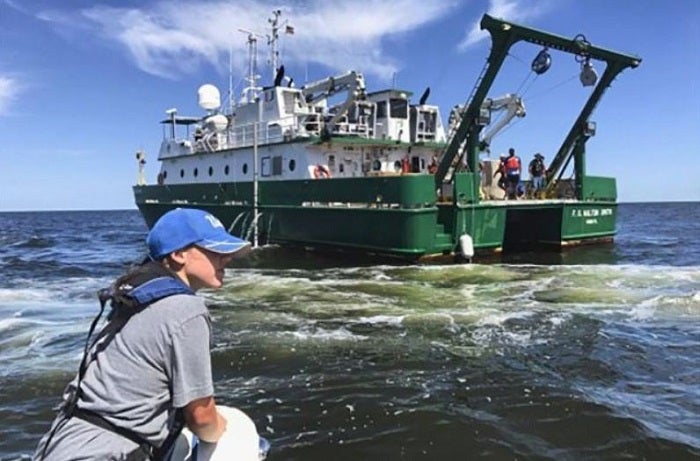 UCLA students conduct oceanic research in the Gulf of Mexico. A student wearing a UCLA hat on water, with a view of a boat in the background.