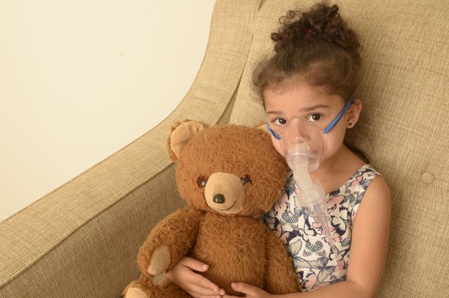 A young girl wearing an oxygen mask, holding a teddy bear.
