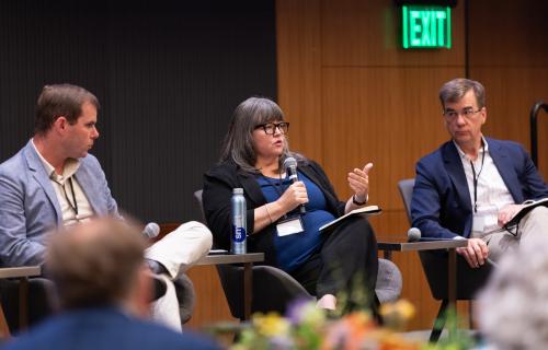 A woman sitting while she talks into a microphone and two people watch on both sides of her.